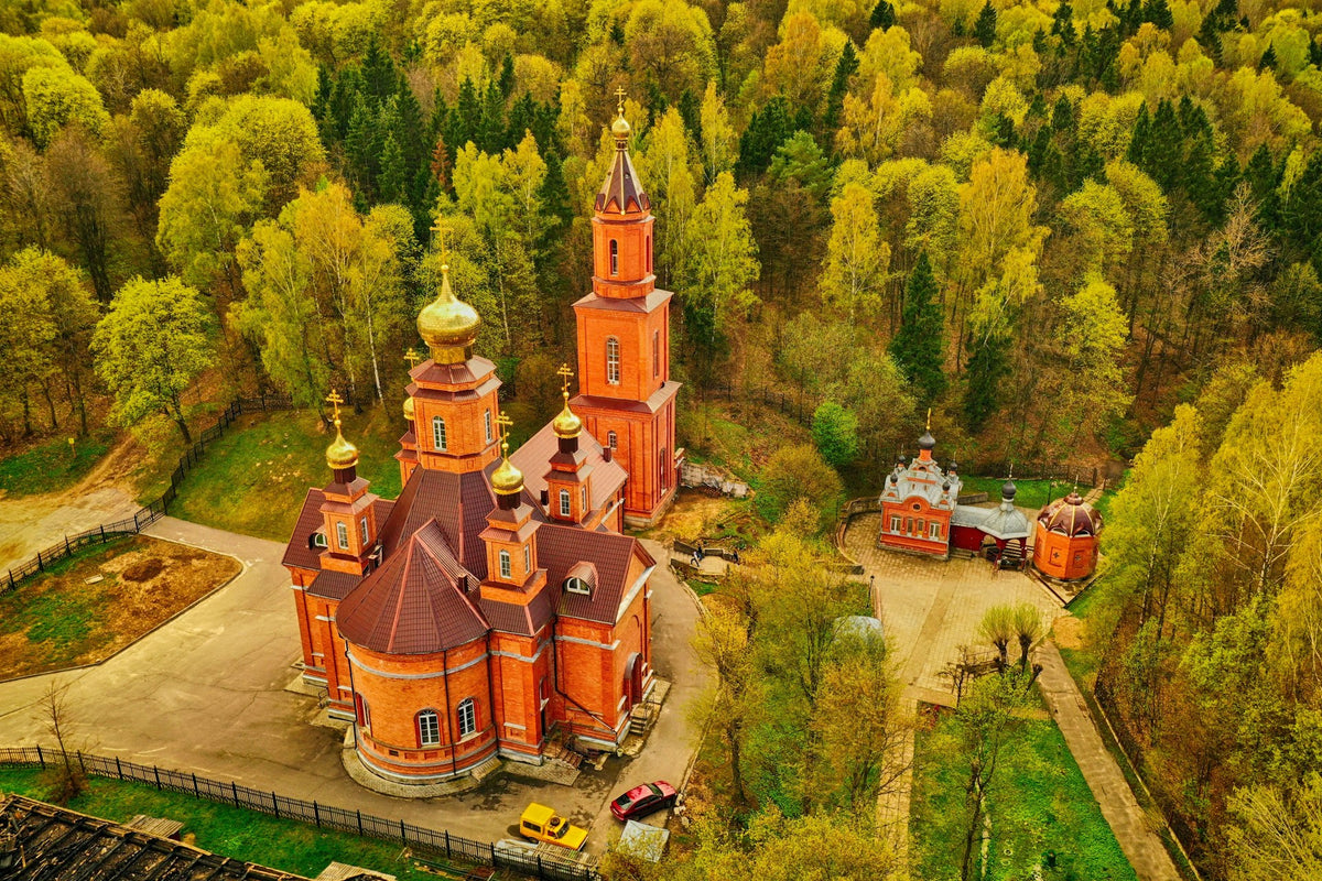 Aerial view of a red brick Orthodox church in Belarus with golden domes surrounded by a dense yellow-green forest.