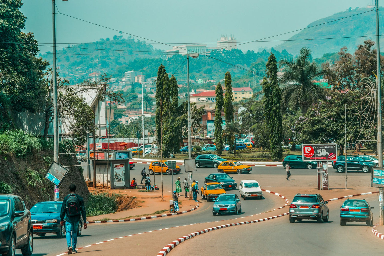 City street in Cameroon with vehicles and pedestrians, surrounded by greenery and hills.