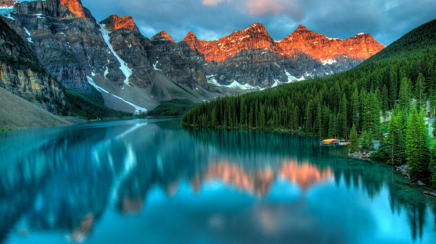 Rocky mountains with light patches of snow are illuminated by warm sunlight casting a soft golden glow, reflected in a shimmering lake and surrounded by a dense forest of green pine trees.