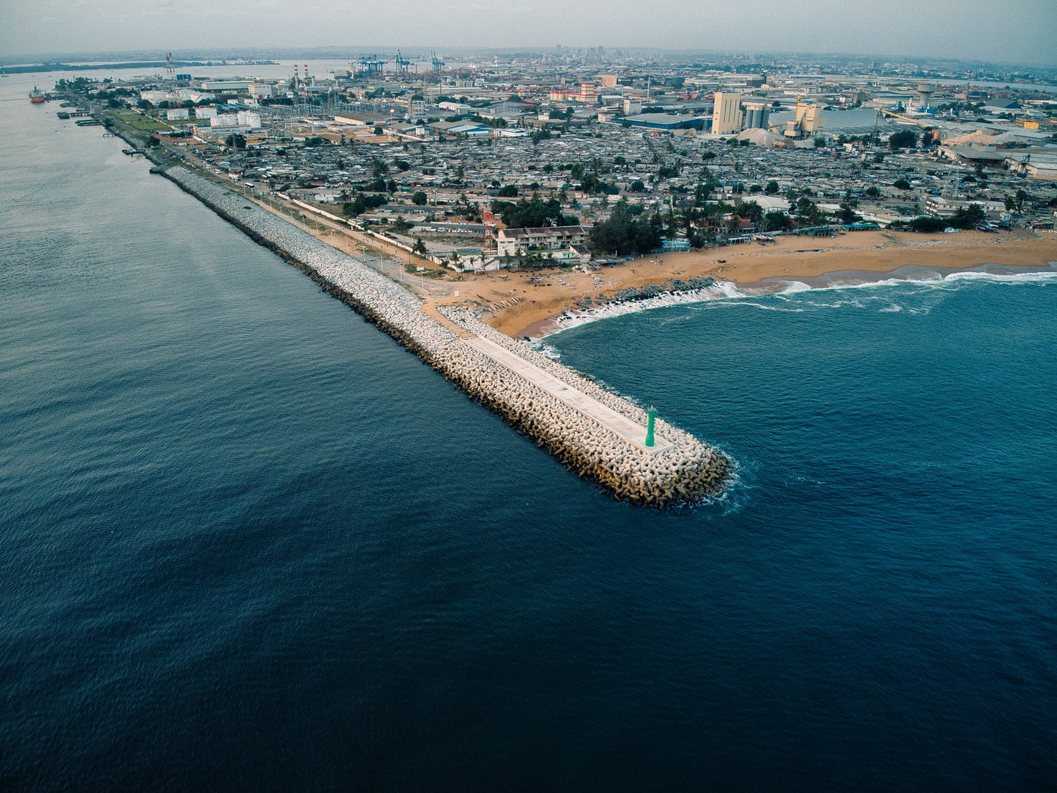 Aerial view of a coastal city in Côte d’Ivoire with a long rocky breakwater extending into deep blue ocean, adjacent to a sandy beach and dense urban area under a hazy sky.