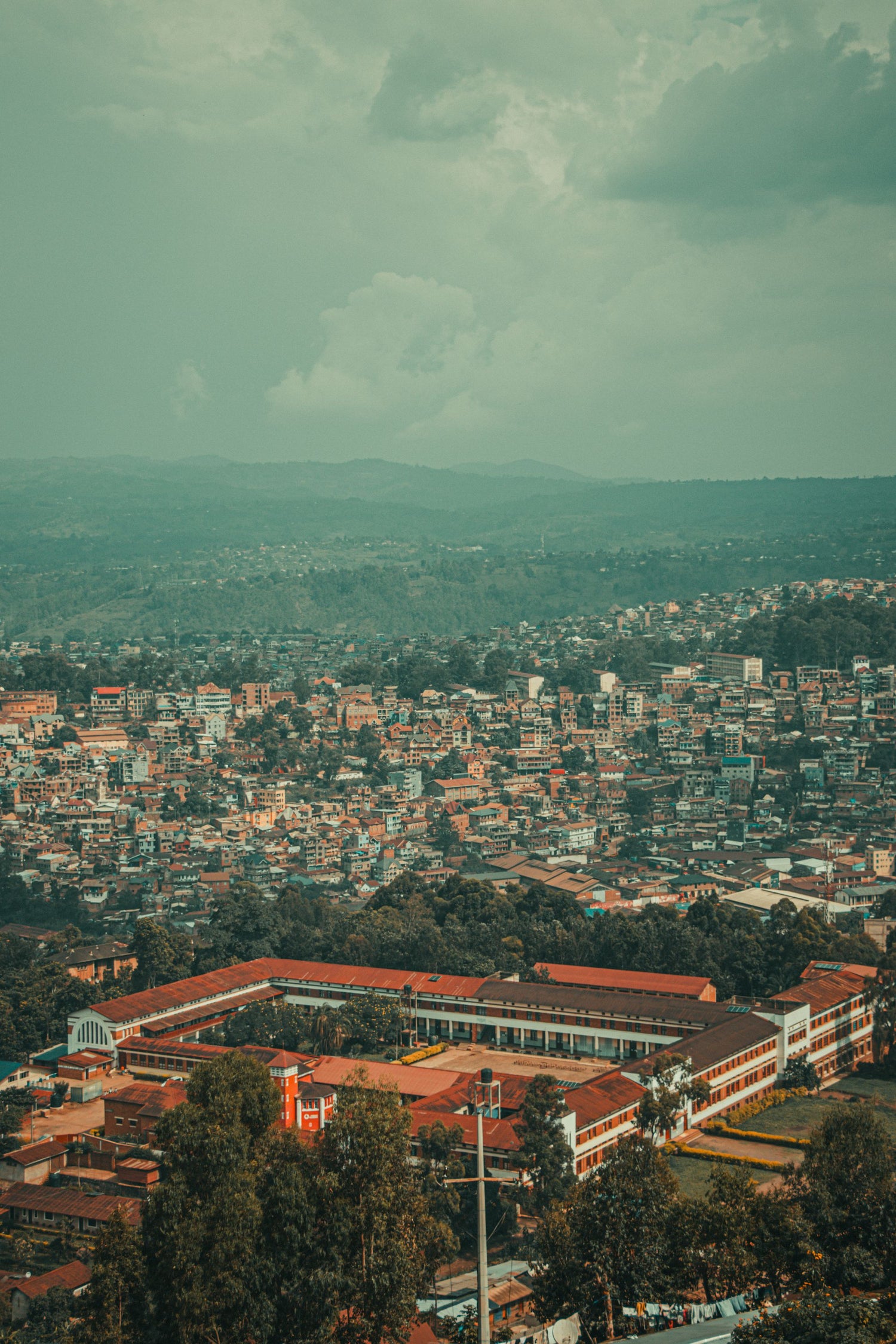 A high-angle view of a dense city in Democratic Republic of the Congo, spread across rolling hills under a cloudy sky, with a large building complex in the foreground surrounded by trees.