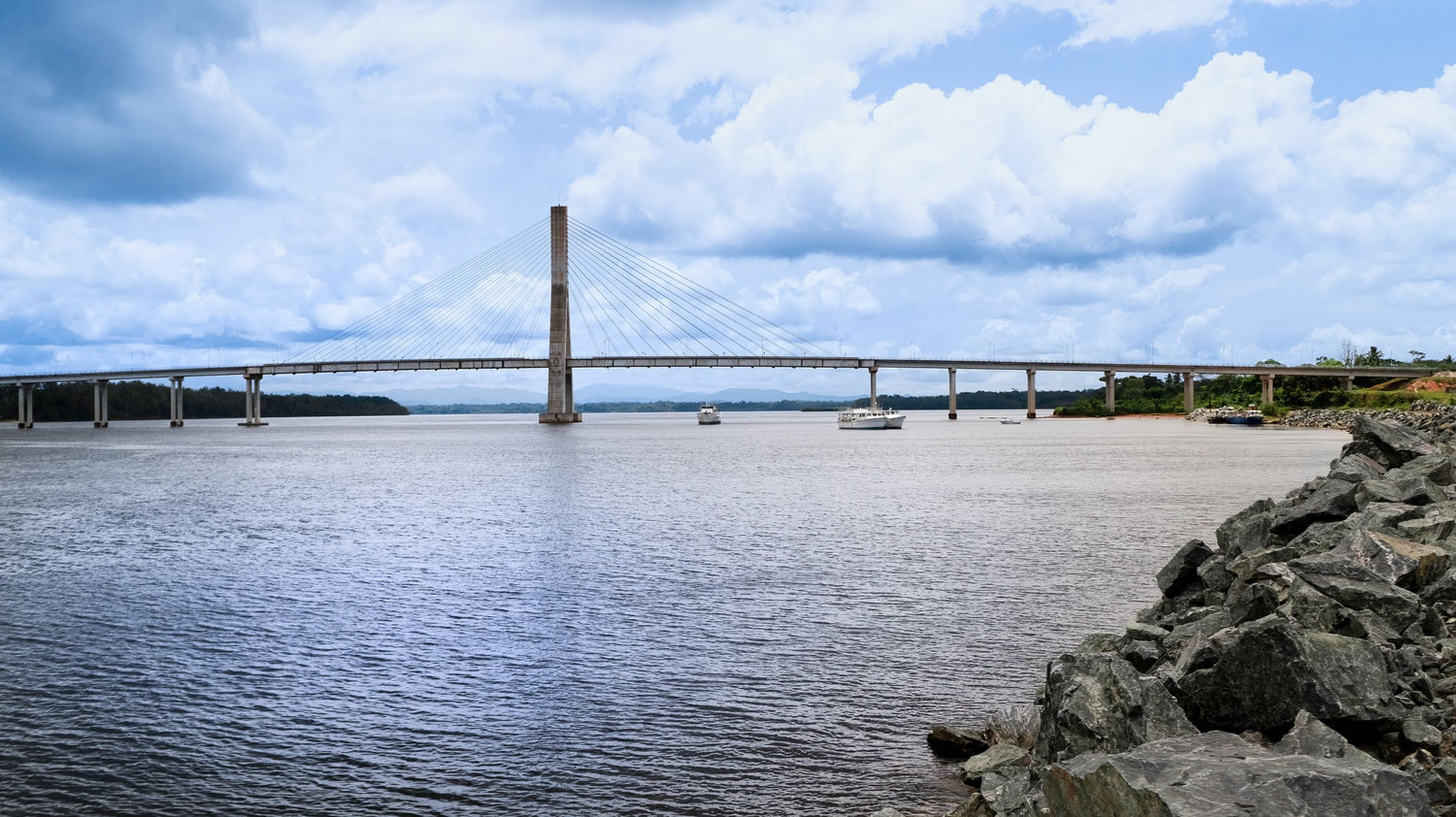 A wide river scene in Equatorial Guinea with a long cable-stayed bridge spanning the water under a blue sky with large white clouds. Rocky shoreline is visible in the foreground, and a few small boats appear on the river.