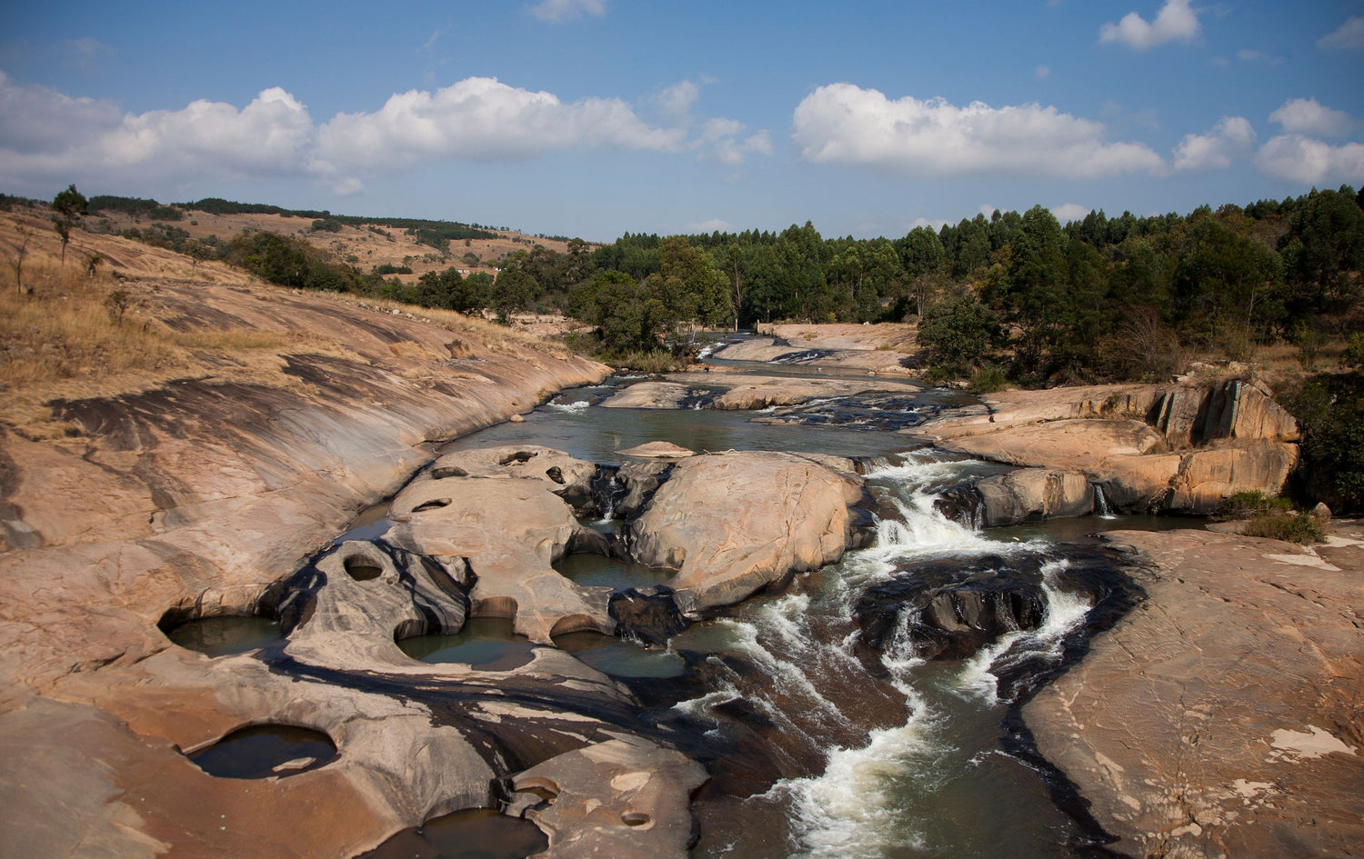A scenic landscape photograph of a rocky river in Eswatini with small cascades flowing over tan and brown stone formations, surrounded by green trees under a blue sky with white clouds.