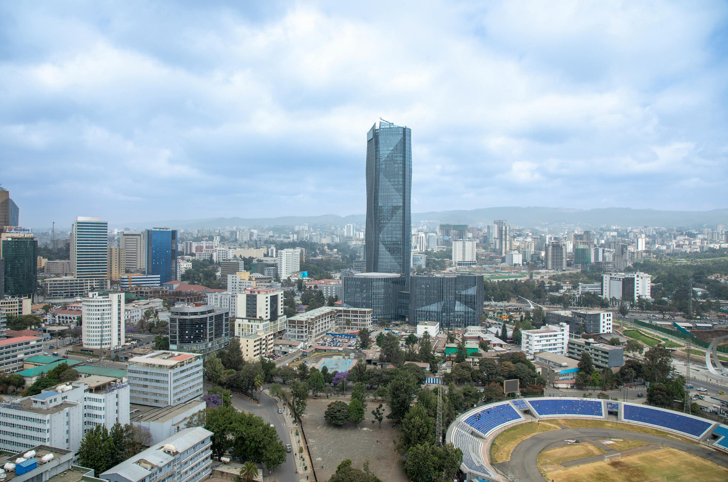Cityscape of Addis Ababa with modern buildings and a sports stadium under a blue sky with a mountain range in the background.