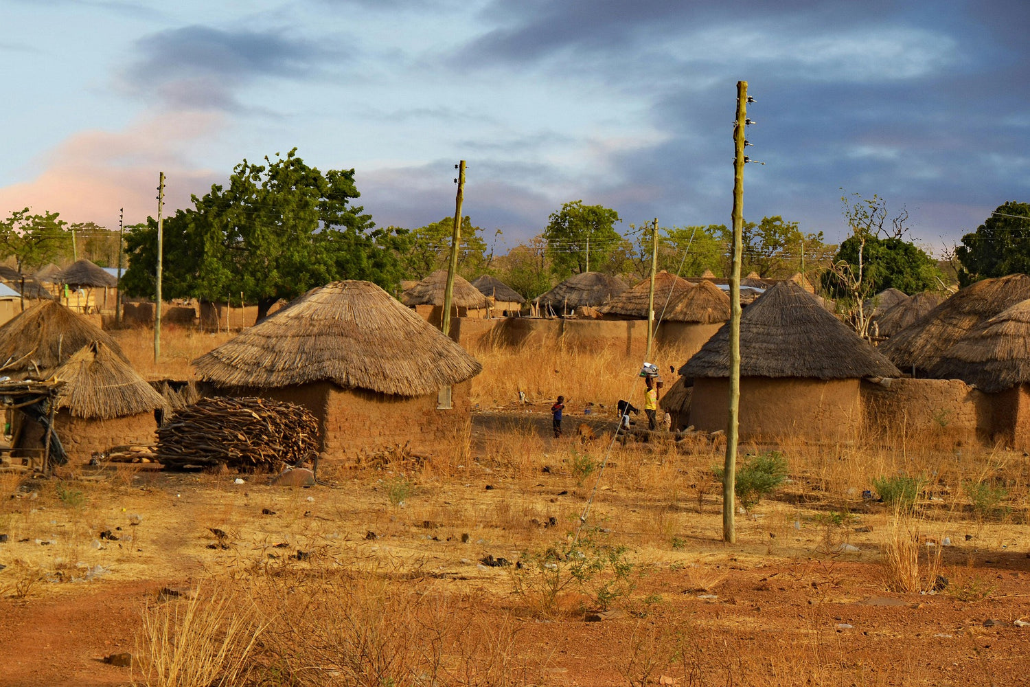 A rural village scene in Ghana with several thatched-roof huts, dry grass, trees, utility poles, and a cloudy blue sky.