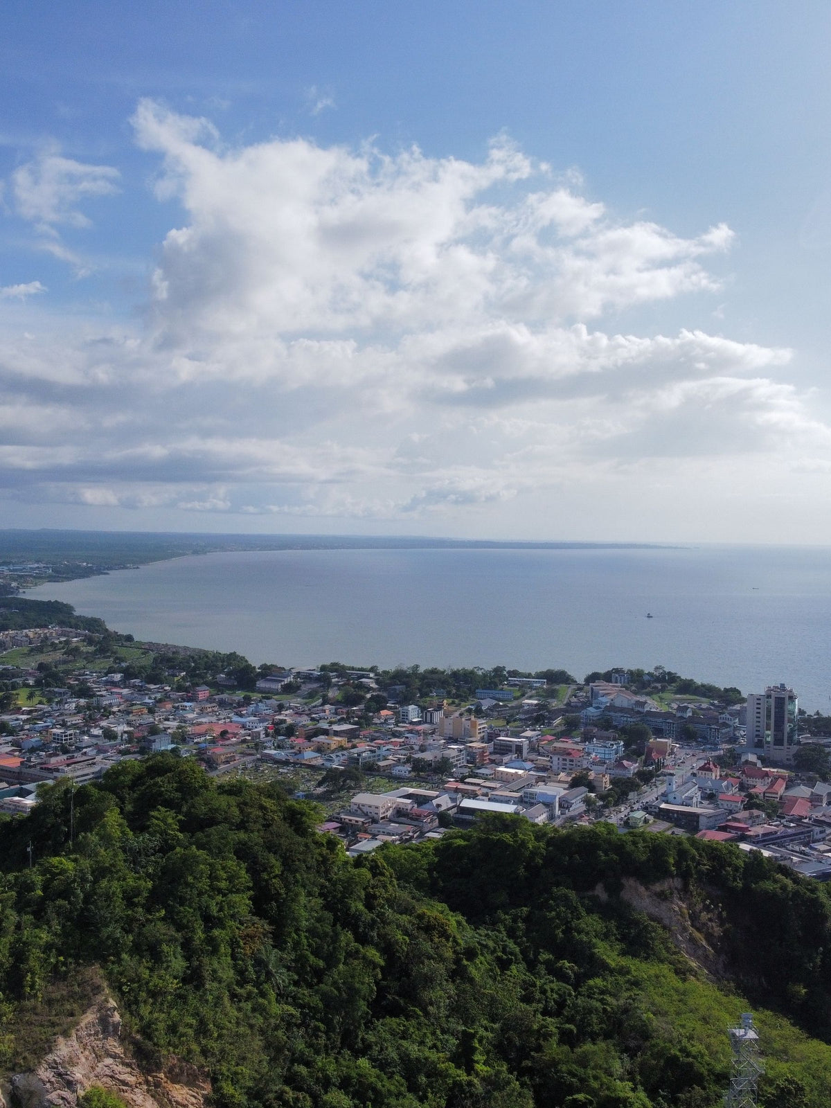A scenic high-angle view of a coastal city in Guyana nestled between lush green hills and a large body of water under a blue sky with white clouds.