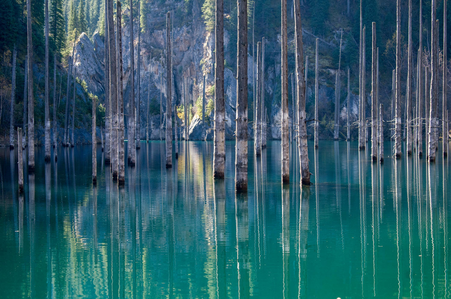 Teal water of Lake Kaindy in Kazakhstan, with submerged tree trunks and mountains in the background.