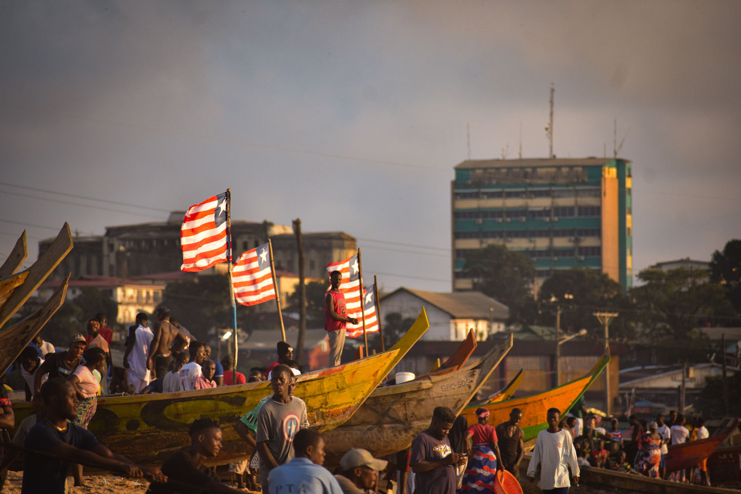 A bustling waterfront scene with many people gathered around traditional wooden boats on a dock, several boats flying red and white striped flags, with buildings and a tall blue-green tower in the background under a hazy sky.