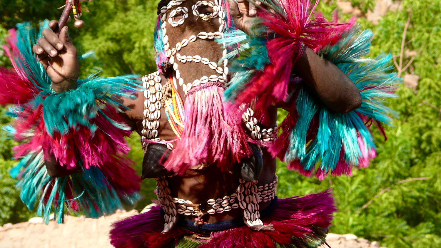 A person in Mali wearing a highly decorative costume with bright pink, teal, and magenta feathered or fringed elements, shell beadwork, and a dark patterned garment against a blurred green natural background.