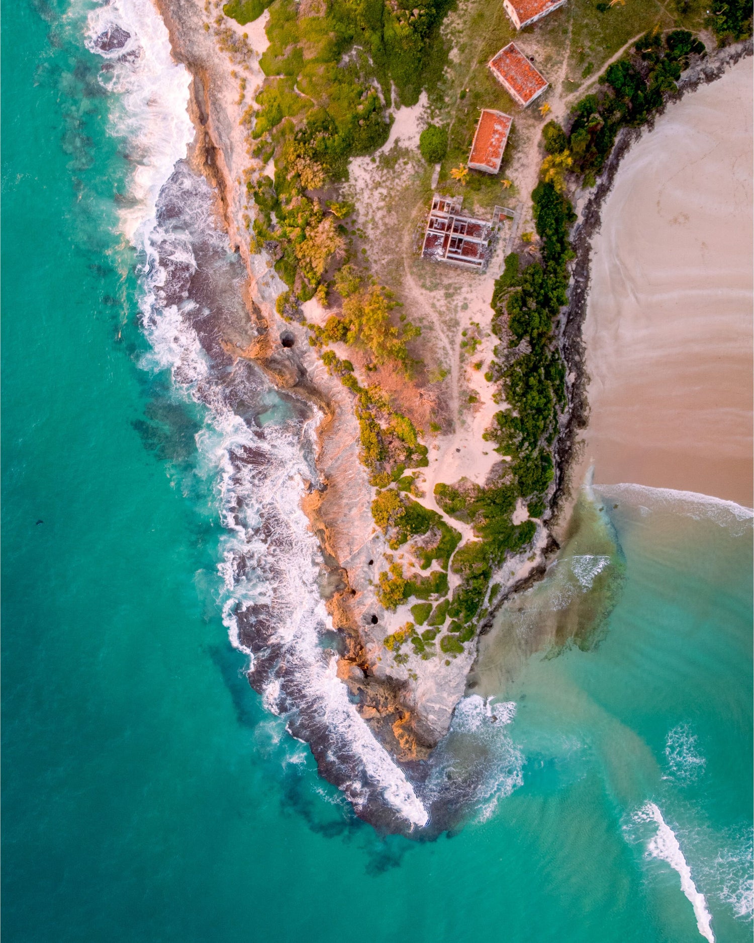 Aerial view of a coastal peninsula in Mozambique with turquoise ocean water, white surf, sandy beach, green vegetation, rocky shoreline, and a few buildings with red roofs.