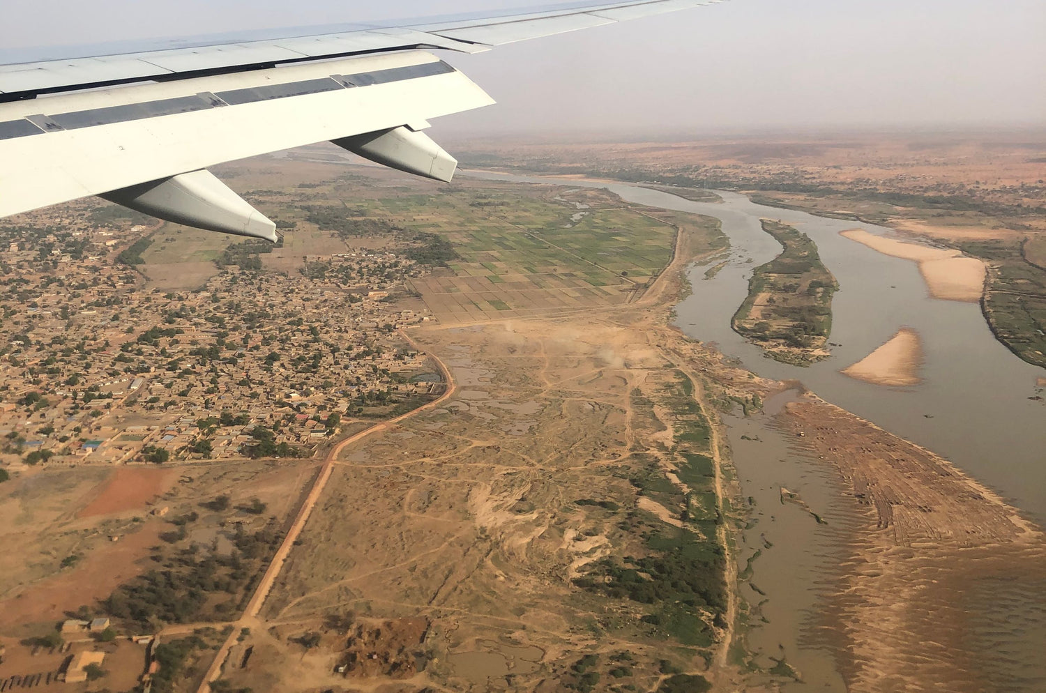 Aerial view of Niger from an airplane window showing a river winding through a dry, sparsely populated landscape below, with part of the aircraft wing visible in the upper left.