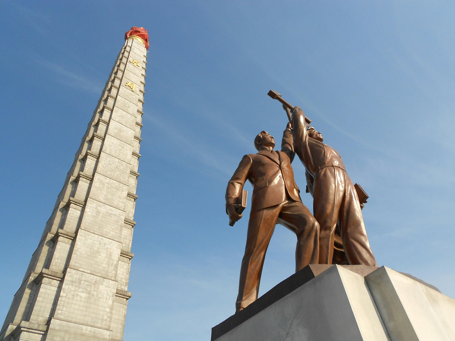 Statue of two figures with a tall pyramid-like structure against a blue sky, North Korea.
