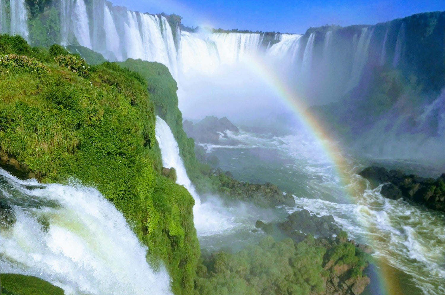 Waterfalls with rapids at the base, greenery, and a misty rainbow.