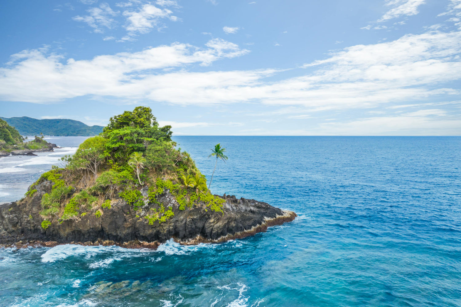 Small island in Samoa with lush greenery surrounded by clear blue water and a partly cloudy sky.