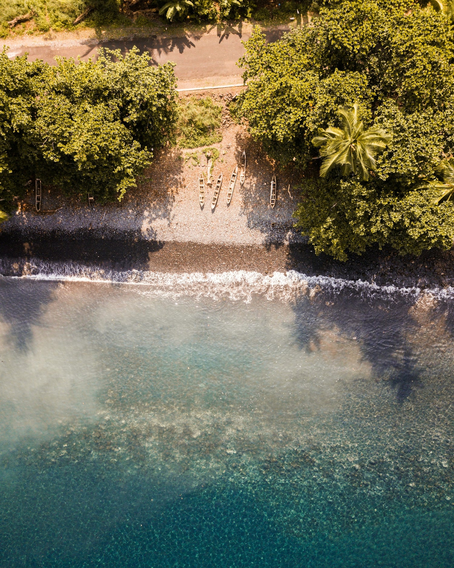 Aerial view of a tropical coastline in São Tomé with clear turquoise water, a dark pebble beach, dense green trees, and a narrow road running along the shore.