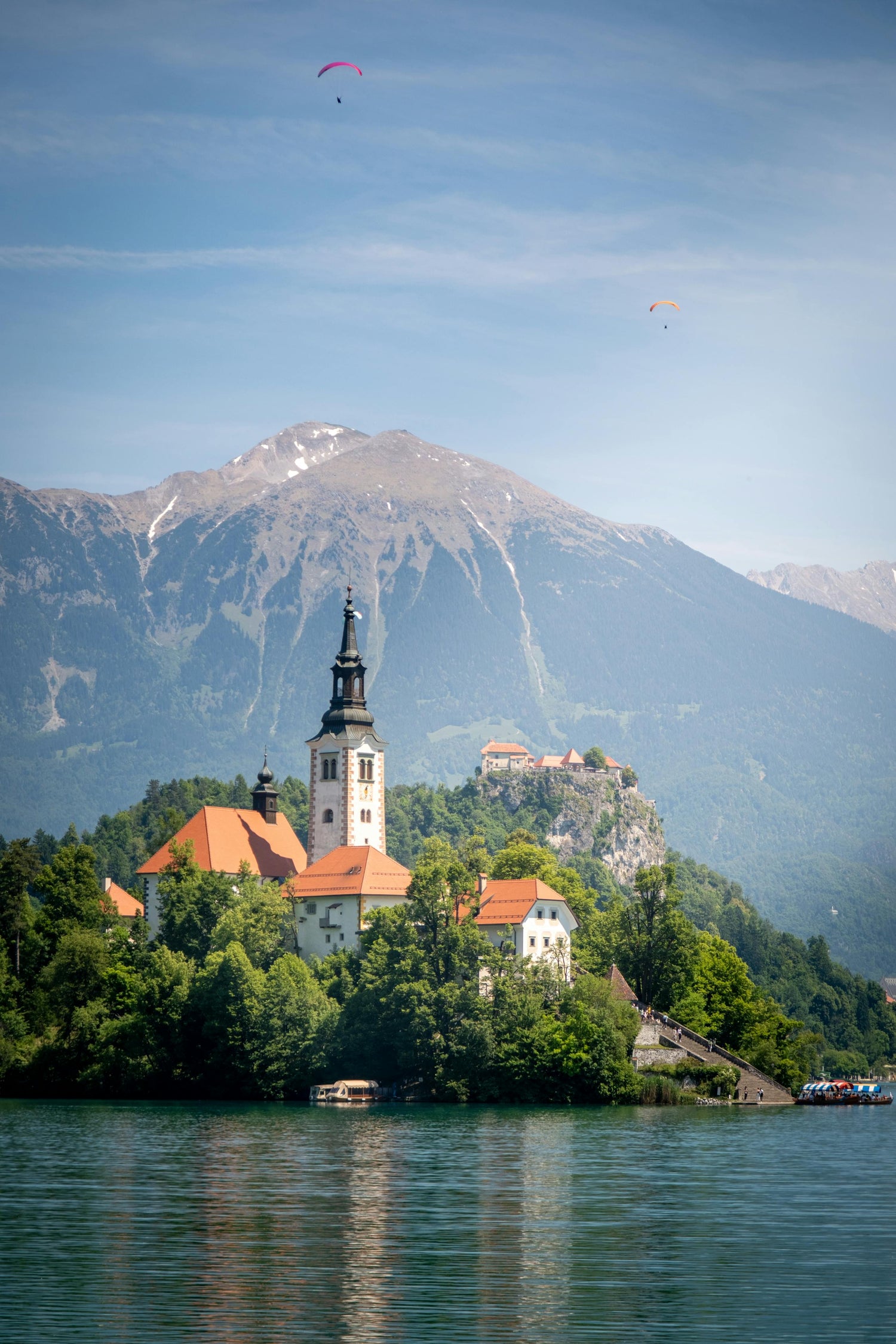 Small island in Slovenia with a church and castle surrounded by water and mountains, with two paragliders in the sky.