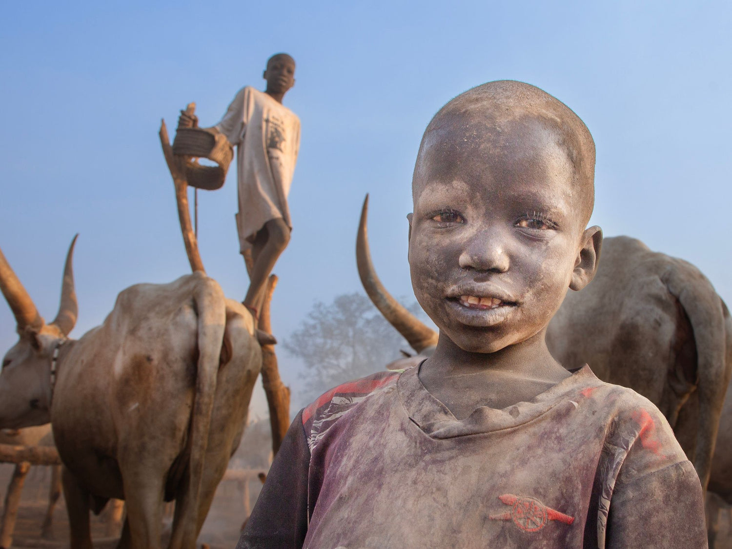 A young child from South Sudan in the foreground with face and clothing covered in dust, standing outdoors near oxen and a person in the background under a blue sky.