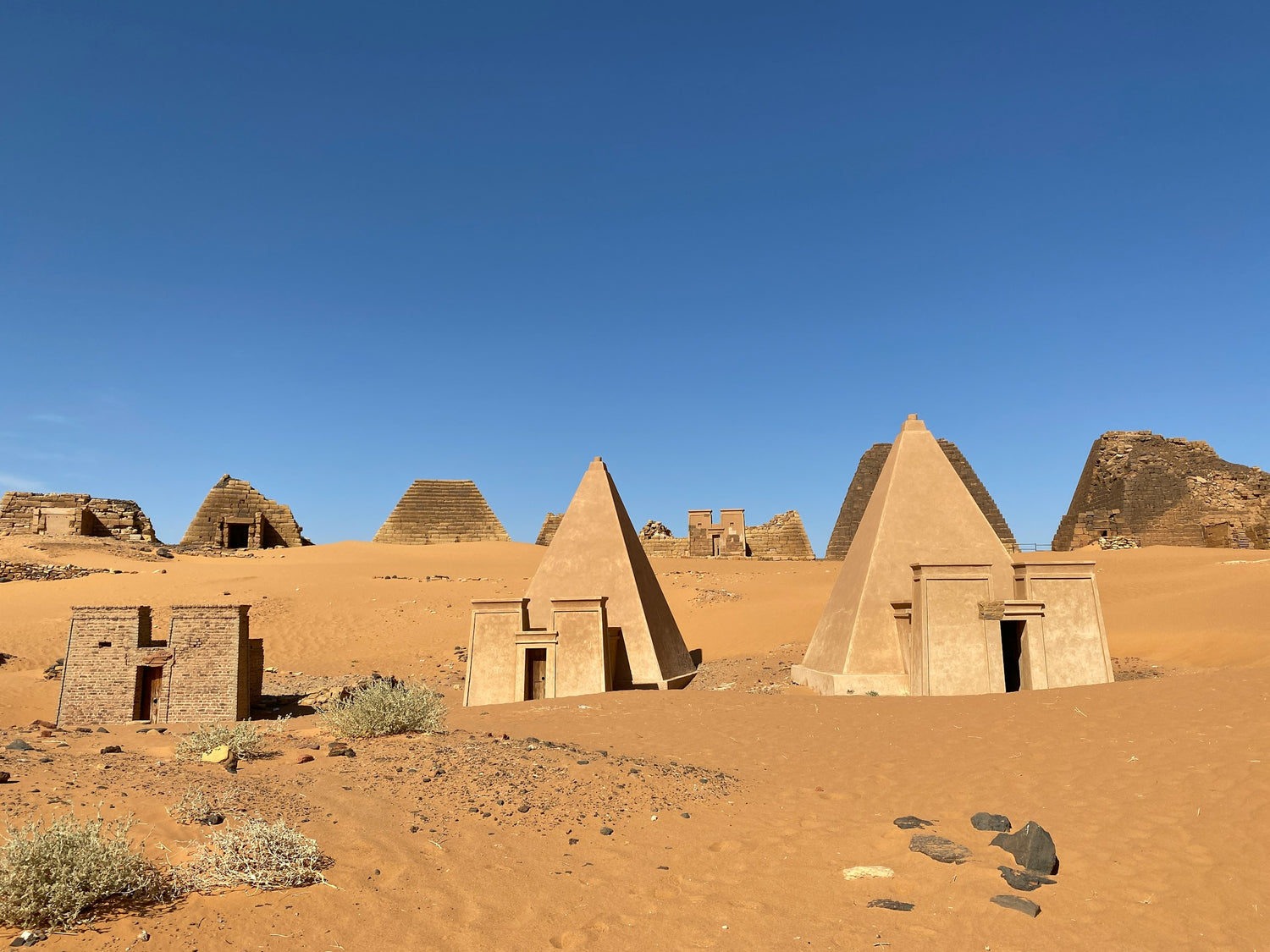 A desert landscape with several small beige pyramid-like structures and rectangular buildings built into sandy dunes under a clear blue sky.