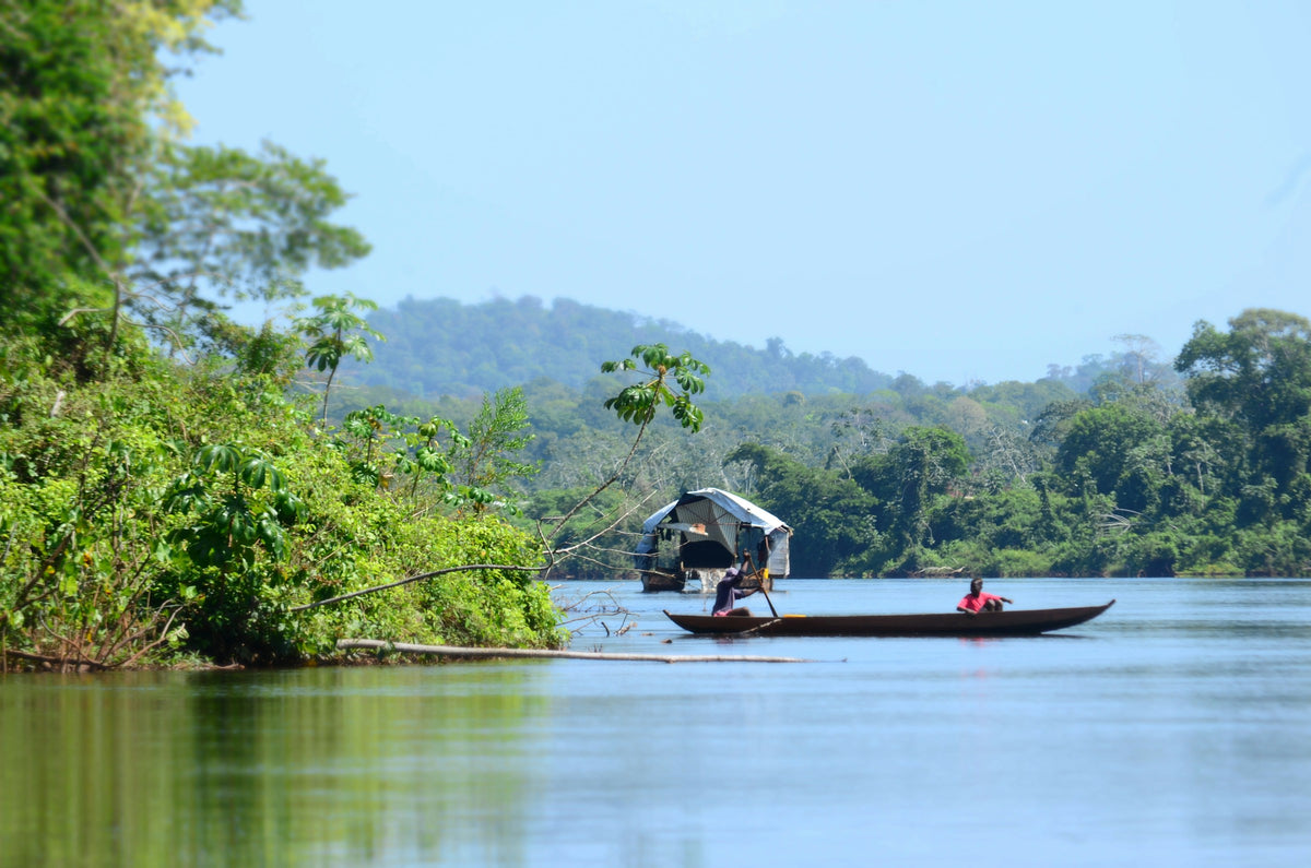 A scenic landscape photograph in Suriname of a river with lush green forest on the banks, a small canopy-covered boat, and a person in a long canoe in the foreground under a clear blue sky.