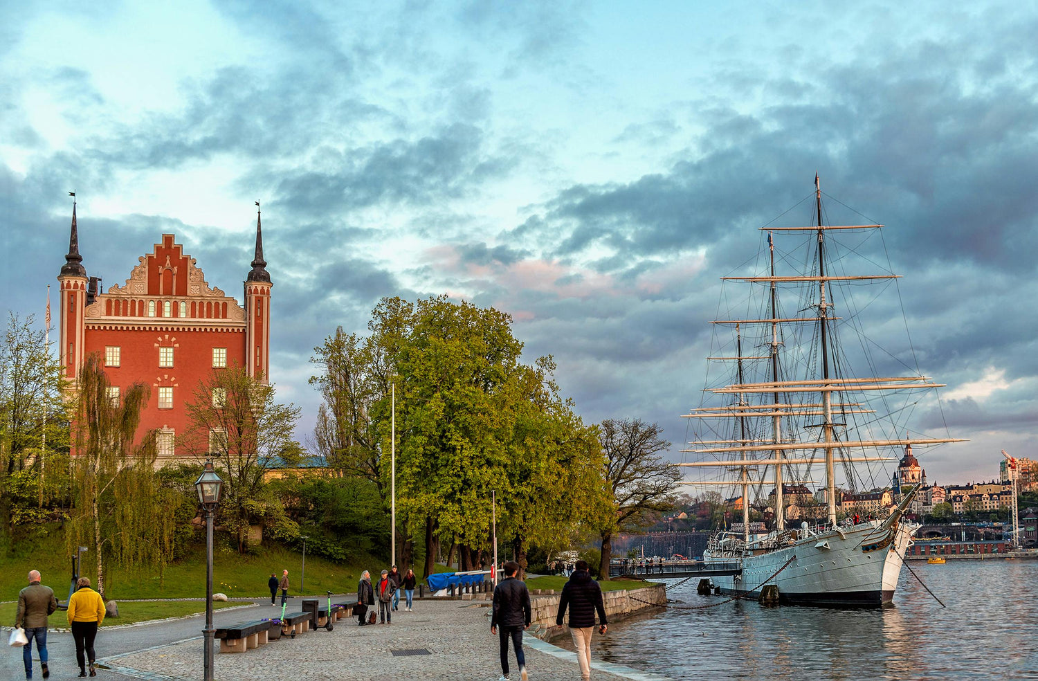 Cityscape in Sweden with a red brick building, tall ship, and people walking along a waterfront.