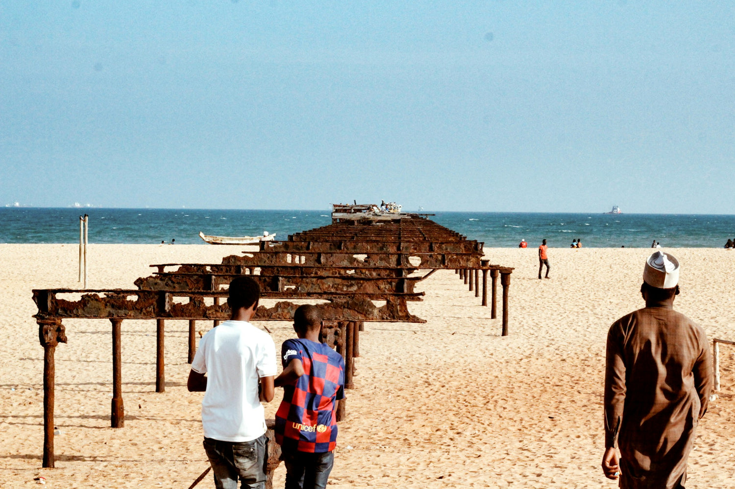 A wide beach scene in Togo with a large rusted metal structure extending into the ocean, with several people walking on the sand under a clear blue sky.