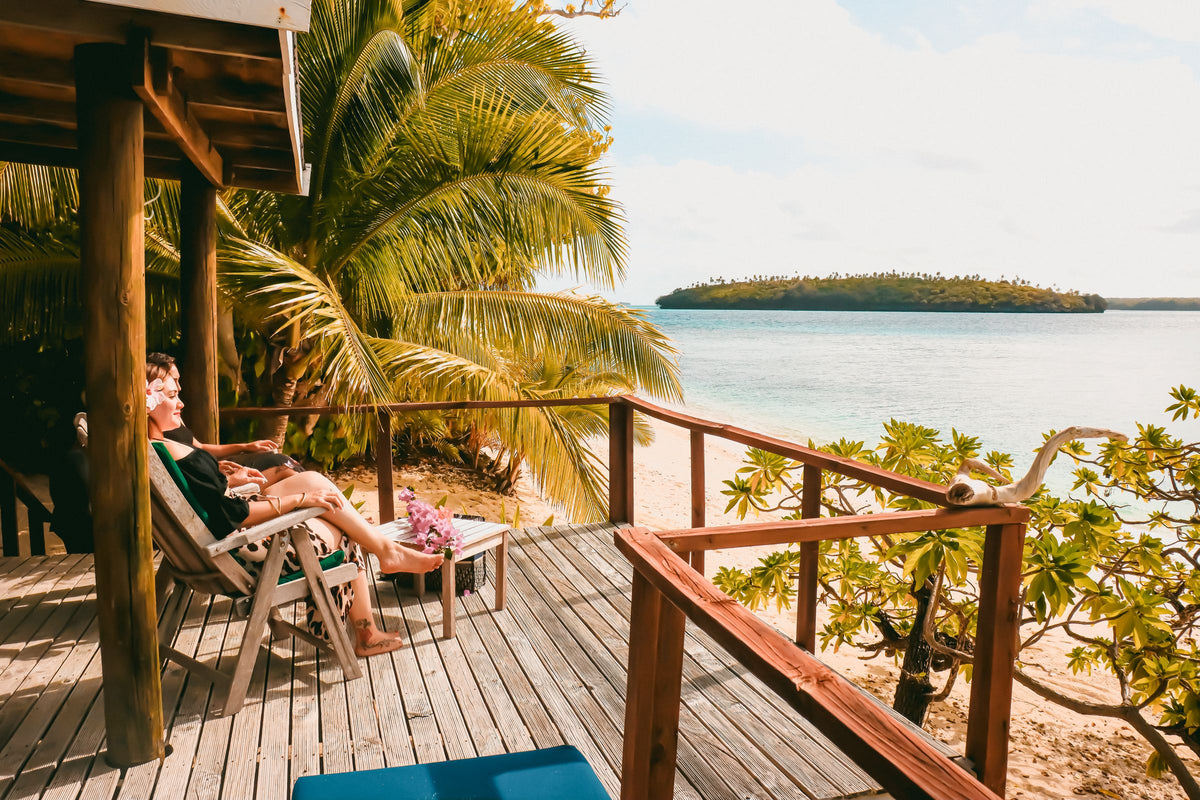 A couple on lounge chairs on a wooden beachfront deck in Tonga, overlooking a tropical ocean, palm trees, and a sandy shoreline. 