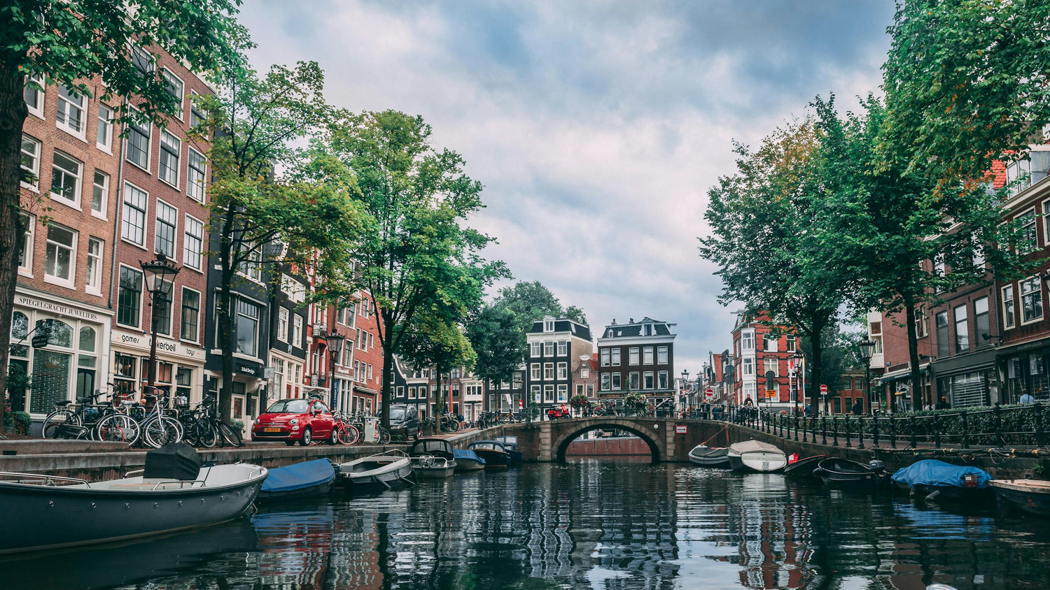 Canal scene in the Netherlands, with reflective water, blue and white boats, old European buildings, and luscious green trees.